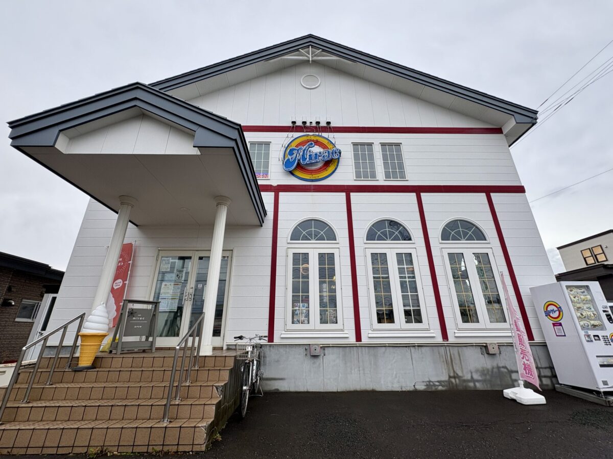 網走市の「カフェ ミラク（Mirac）」の店舗外観。白い外壁に赤いフレームの装飾が施された2階建ての建物。入り口にはソフトクリームのオブジェがあり、壁面には青・赤・黄色の円形ネオン看板が掲げられている。右側には自動販売機も設置されている。
The exterior of "Cafe Mirac" in Abashiri, Hokkaido. A white two-story building with distinctive red trim. The entrance features a soft-serve ice cream display, and a retro-style neon sign is mounted on the wall. A vending machine is visible to the right.