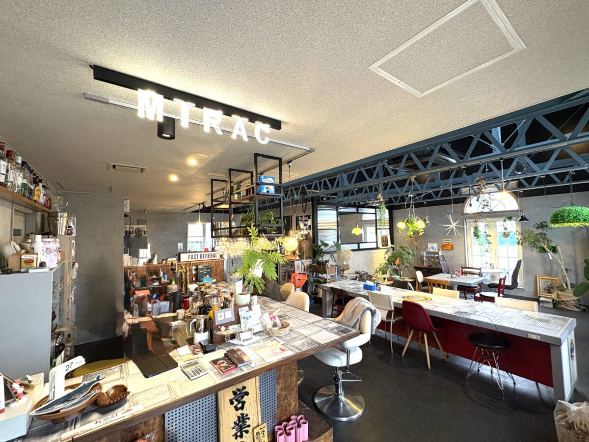 網走市「カフェ ミラク（Mirac）」の店内。手前にはレトロな木製カウンターと、天井には「MIRAC」の文字を象ったおしゃれなネオン照明がある。奥にはインダストリアルな鉄骨天井と、植物がたっぷりと飾られたテーブル席が広がり、レトロモダンな雰囲気が漂う。
Interior view of "Cafe Mirac" in Abashiri. A vintage wooden counter is in the foreground, with unique "MIRAC" neon sign lighting on the ceiling. The background features an industrial steel truss ceiling and table seating surrounded by abundant green plants, creating a retro-modern atmosphere.