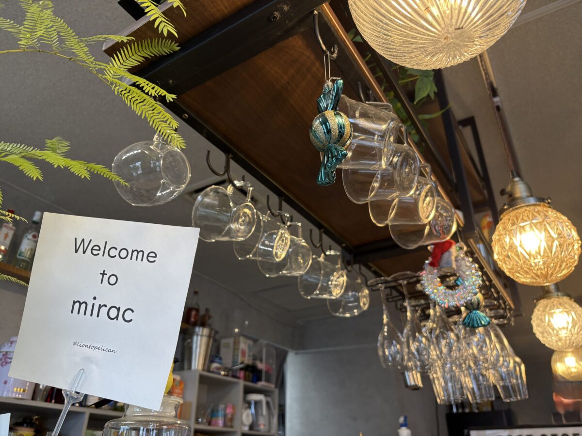 網走「カフェ ミラク（Mirac）」のカウンター上部。ハンギングラックに整然と吊り下げられた透明なティーカップやワイングラスが並んでいる。キャンディ型のオーナメントや温かい光のペンダントライトが飾られ、手前には「Welcome to mirac」と書かれたメッセージカードが置かれている。
A stylish counter display at "Cafe Mirac" in Abashiri. Transparent tea cups and wine glasses are neatly hanging from a wooden overhead rack. The area is decorated with festive candy-shaped ornaments and warm glowing pendant lights, with a "Welcome to mirac" card in the foreground.