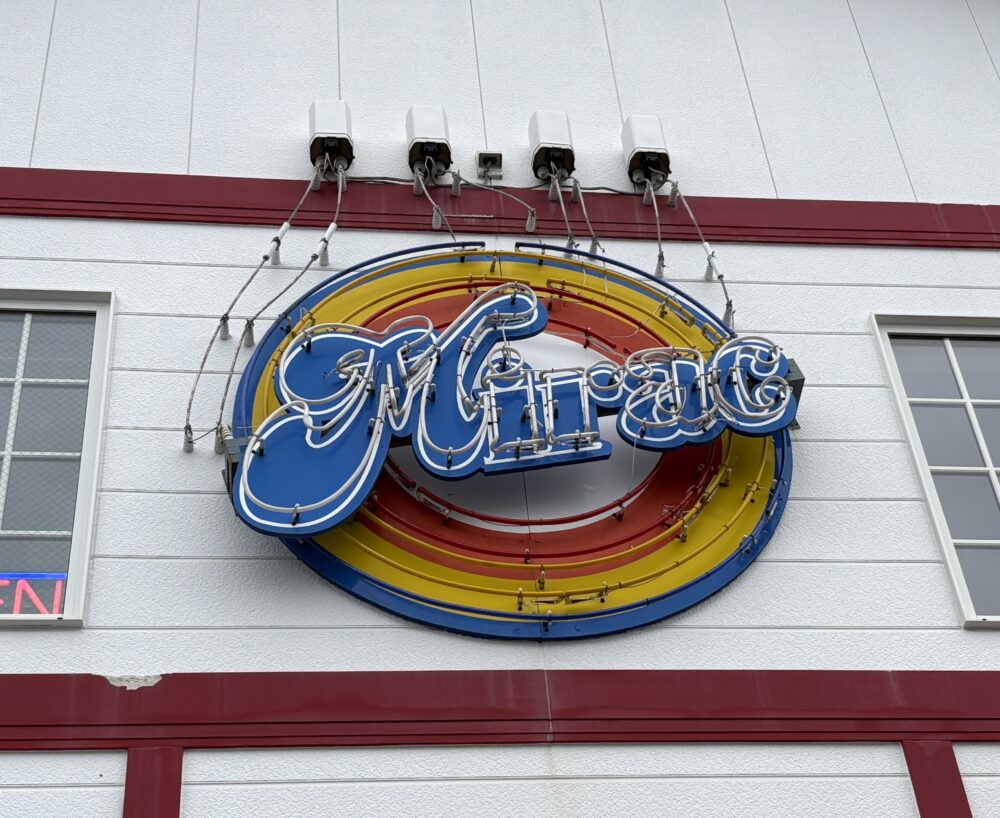 「カフェ ミラク（Mirac）」の店舗壁面にあるネオンサインのアップ。青色のベースに白の筆記体で「Mirac」と書かれ、背後には黄色と赤の同心円状のネオン管が配置されたレトロでポップなデザイン。 A close-up of the neon sign for "Cafe Mirac" in Abashiri. The sign features "Mirac" in white cursive script over a blue background, accented with yellow and red concentric neon rings, giving it a retro American diner feel.