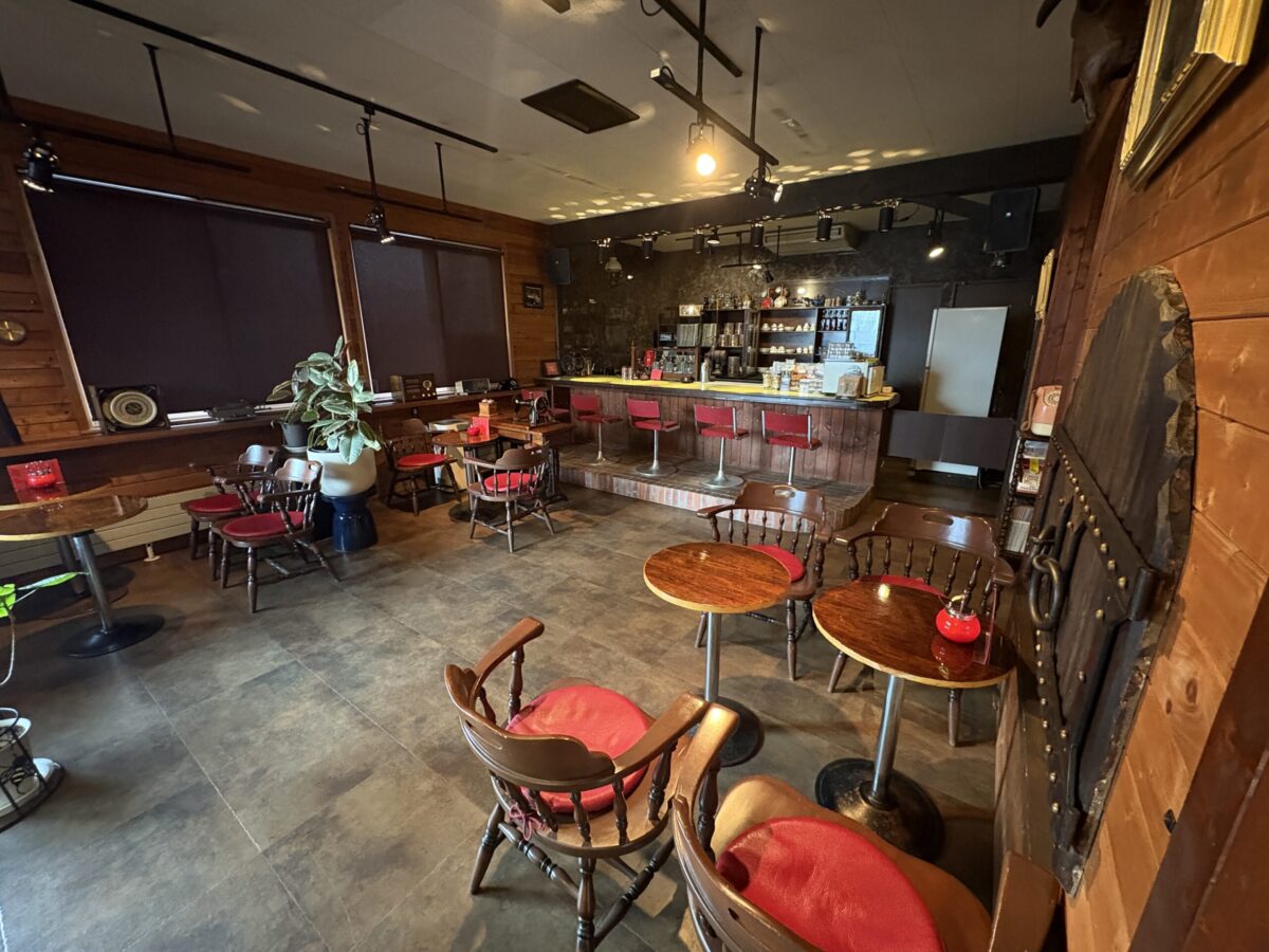 網走「珈琲屋 デリカップ」の店内風景。赤い座面が印象的な木製の椅子と円形テーブルが並び、奥にはジャズが聞こえてきそうなシックなカウンター席がある、広々としたくつろぎの空間。
The interior seating area of "Coffee-ya Delicup" in Abashiri, showing classic wooden chairs with red cushions and a retro-style counter bar.