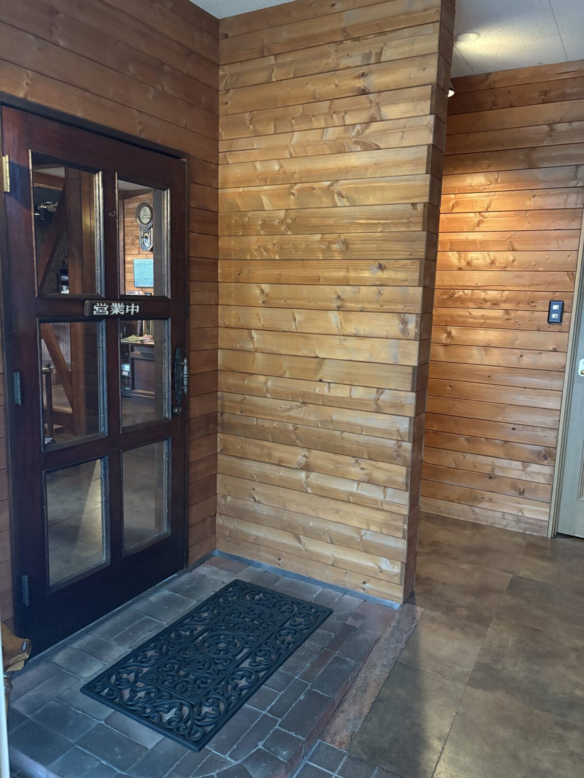 網走の「珈琲屋 デリカップ」の店内入り口。温かみのある木目調の壁と、重厚な木製ドアが特徴的。ドアには「営業中」の看板が掲げられ、落ち着いた喫茶店の雰囲気が伝わるエントランス。
The entrance of "Coffee-ya Delicup" in Abashiri, featuring warm wooden walls and a classic wooden door with an "Open" sign, creating a cozy and welcoming atmosphere.