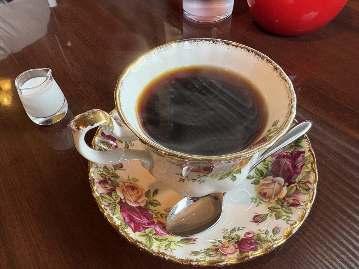 網走の「珈琲屋 デリカップ」で提供される自家焙煎のホットコーヒー。華やかなバラの絵柄が描かれたアンティーク調のカップとソーサー、横には小さなミルクピッチャーが添えられている。
A cup of freshly brewed hot coffee at "Coffee-ya Delicup" in Abashiri, served in an elegant floral teacup and saucer with a small pitcher of milk on the side.