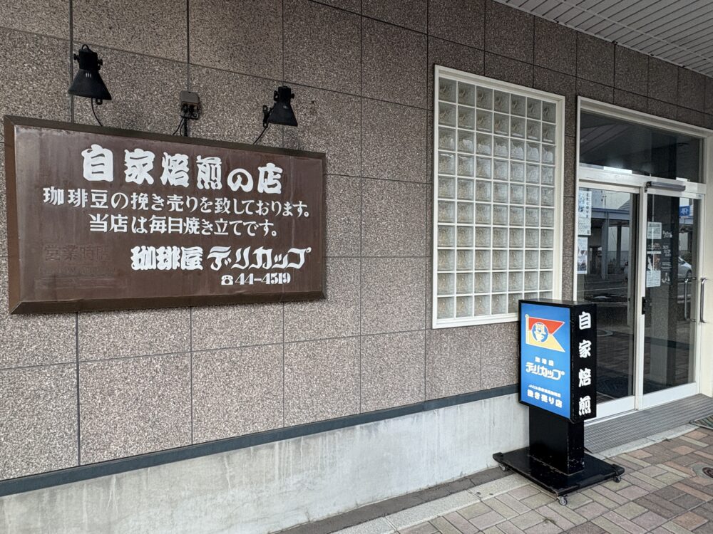 網走市の自家焙煎コーヒー豆販売店「珈琲屋 デリカップ」の外観。茶色の大きな看板には「毎日焼き立て」の文字があり、青い立て看板が入り口に置かれている。 Exterior of "Coffee-ya Delicup," a home-roastery in Abashiri, featuring a large brown sign and a blue standing signboard at the entrance.