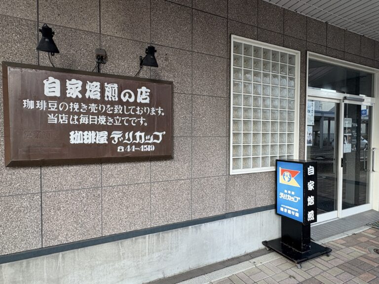 網走市の自家焙煎コーヒー豆販売店「珈琲屋 デリカップ」の外観。茶色の大きな看板には「毎日焼き立て」の文字があり、青い立て看板が入り口に置かれている。 Exterior of "Coffee-ya Delicup," a home-roastery in Abashiri, featuring a large brown sign and a blue standing signboard at the entrance.