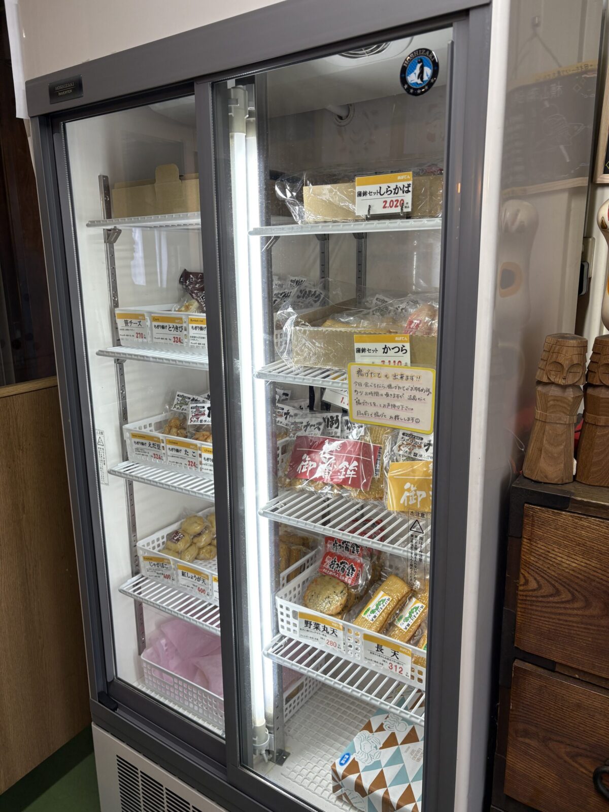 網走「横山蒲鉾店」の店内にある大型の冷蔵ショーケース。地方発送も可能な贈答用セットや、自宅用のお土産にぴったりな多種類のかまぼこが整然と並んでいる。
A large refrigerated display case inside Yokoyama Kamaboko in Abashiri, showcasing various fish cake gift sets and locally made seafood products ready for purchase or shipping.