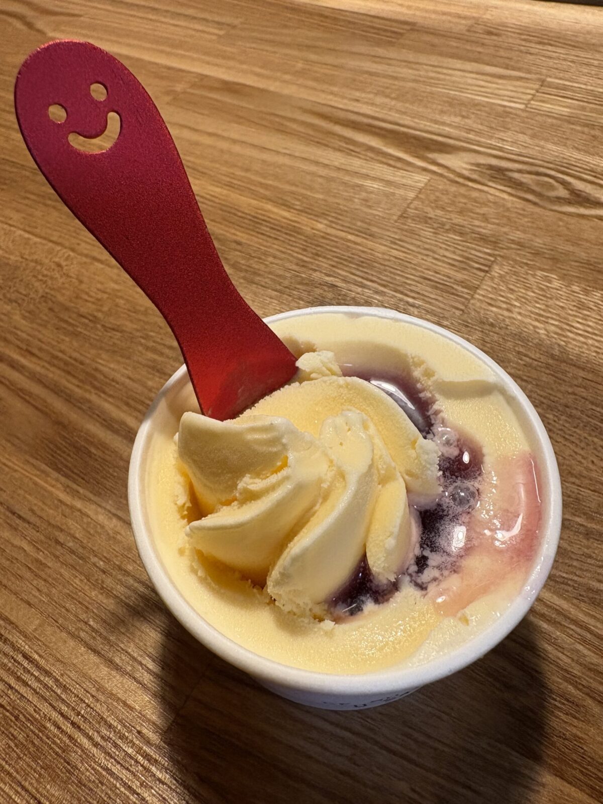 網走「フラミンゴ」の食後のデザート。カップに入ったバニラアイスに、鮮やかなベリー系のソースが添えられています。スマイルマークが切り抜かれた真っ赤なスプーンが差し込まれており、木目調のテーブルの上で可愛らしく盛り付けられています。
A cup of vanilla ice cream topped with berry sauce, served at "Flamingo" in Abashiri. A bright red spoon with a carved-out smiley face is dipped into the ice cream, all set against a warm wooden table background.