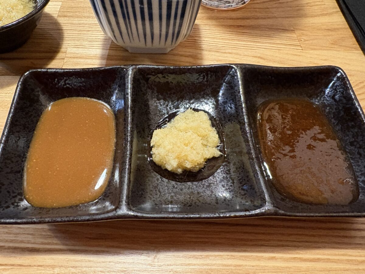 網走「フラミンゴ」のジンギスカンのタレと薬味。黒い3連皿に盛り付けられており、左側には明るい茶色の味噌ベースのタレ、中央にはたっぷりのすりおろし生姜、右側には深みのある色の醤油ベースのタレが入っています。木目調のテーブルの上で、食欲をそそる準備が整った様子です。
A set of three dipping sauces and condiments for Genghis Khan (mutton BBQ) at "Flamingo" in Abashiri. A black rectangular dish features a light brown miso-based sauce on the left, a generous portion of fresh grated ginger in the center, and a rich dark soy-based sauce on the right, all set on a wooden table.