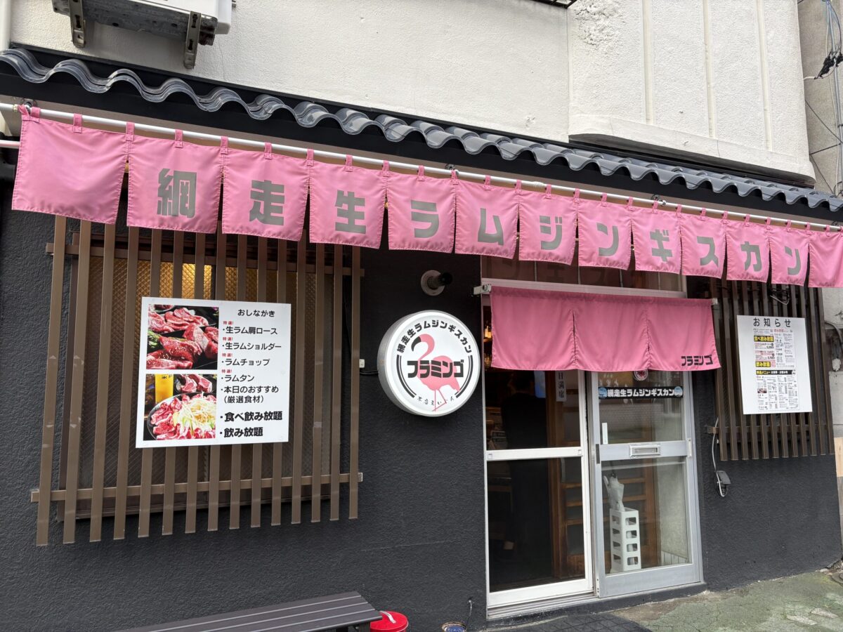 網走のジンギスカン専門店「フラミンゴ」の店舗外観。黒い壁に映える鮮やかなピンク色の暖簾（のれん）と、フラミンゴのイラストが描かれた丸い看板が特徴です。店外の掲示板には「生ラム肩ロース」や「食べ飲み放題」などのメニュー表が掲示されています。
Exterior of "Flamingo," a Genghis Khan (mutton BBQ) restaurant in Abashiri, Hokkaido. The storefront features a striking pink noren curtain and a round sign with a flamingo logo. Menu boards on the wall list specialties like "Raw Lamb Shoulder" and "All-you-can-eat" options
