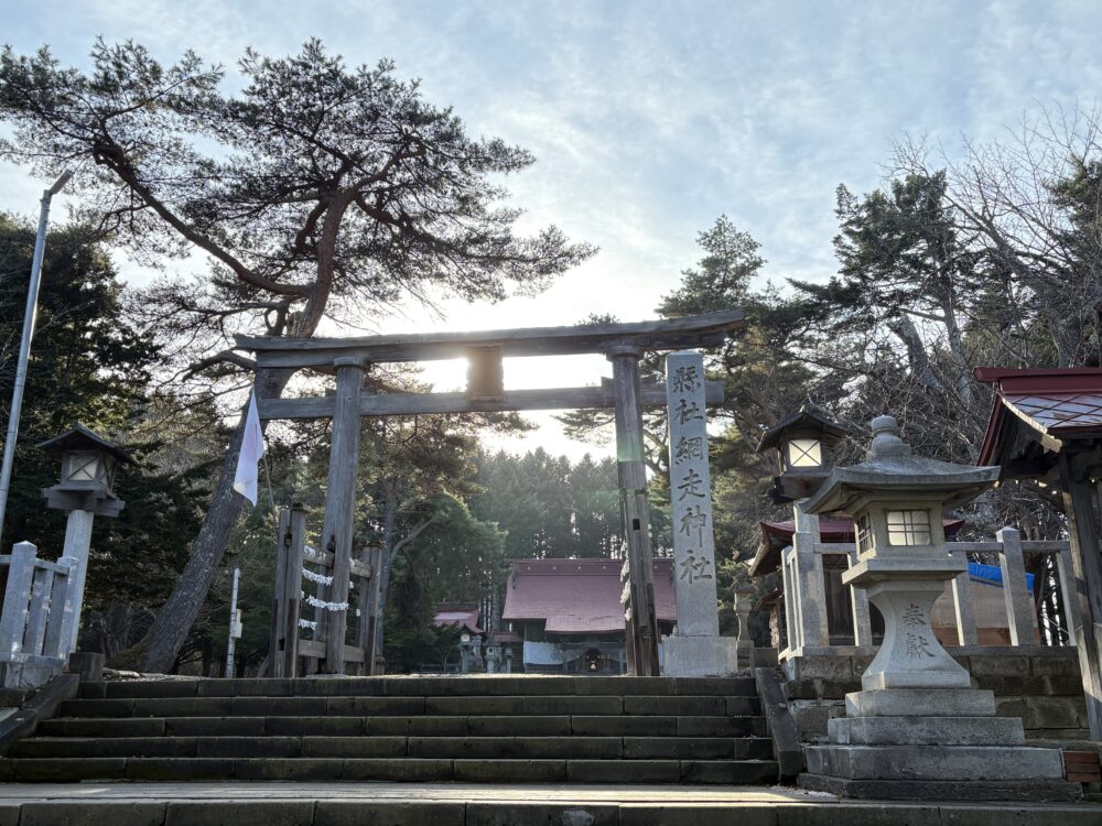 網走神社の入り口に立つ重厚な木造の鳥居と石灯籠。背後から差し込む太陽の光が木々を照らし、神聖な雰囲気が漂う境内の風景。 A majestic wooden Torii gate at the entrance of Abashiri Jinja Shrine, framed by traditional stone lanterns and sunlight filtering through the trees.