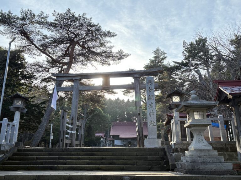 網走神社の入り口に立つ重厚な木造の鳥居と石灯籠。背後から差し込む太陽の光が木々を照らし、神聖な雰囲気が漂う境内の風景。 A majestic wooden Torii gate at the entrance of Abashiri Jinja Shrine, framed by traditional stone lanterns and sunlight filtering through the trees.