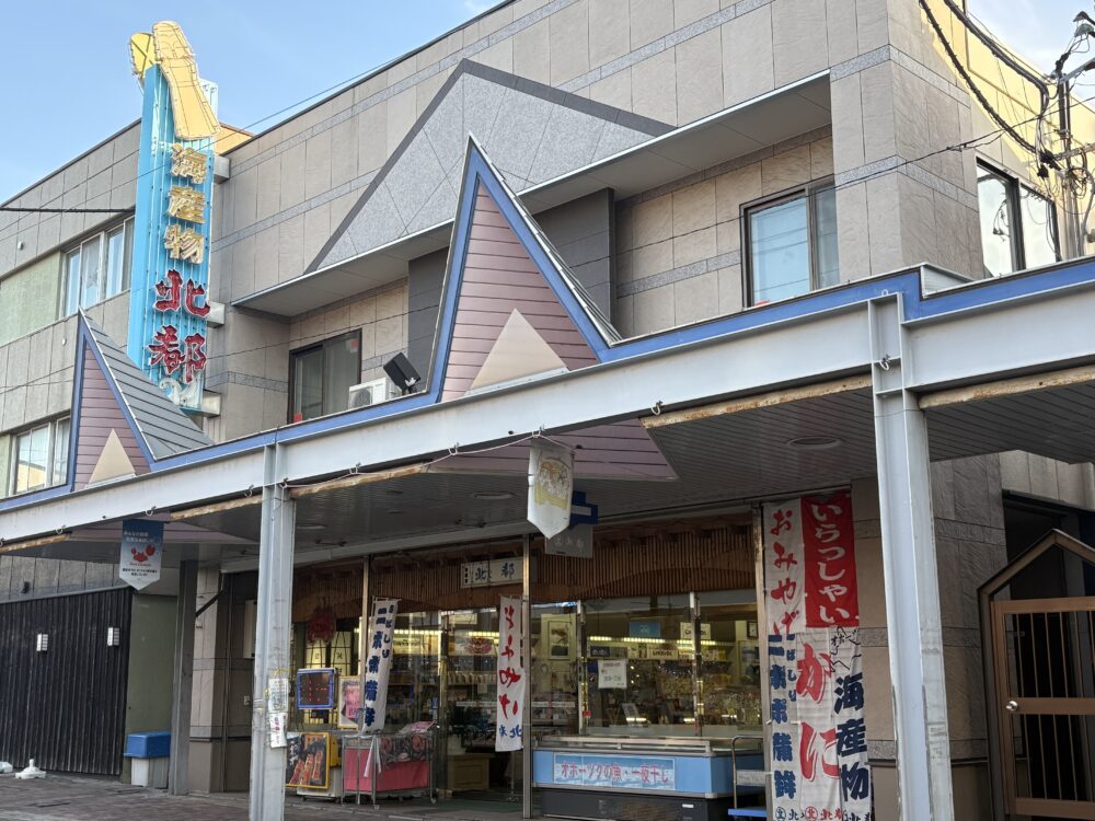 網走市の海産物販売店「丸北北都（まるきたほくと）」の店舗外観。青い看板に白文字で店名が書かれ、入り口付近には「地方発送承ります」の文字やカニのイラストが見える。 Exterior of "Marukitahokuto," a seafood market in Abashiri. The blue sign features the shop name in white, with a crab illustration and signs offering domestic shipping services.