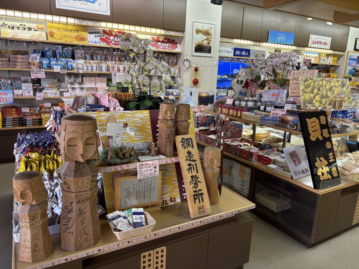 網走「丸北北都」の店内ディスプレイ。手前には大小様々なサイズの「ニポポ人形」の木彫りが並び、その横には「網走刑務所」と書かれた木製の看板が大きく掲げられている。奥の棚にはお菓子や貝柱などの網走・北海道土産が棚いっぱいに並んでおり、この店ならではの品揃えが見て取れる。 Inside Marukitahokuto in Abashiri, featuring a prominent display of various sizes of traditional "Nipopo" wooden dolls in the foreground. Beside them, there is a large wooden sign reading "Abashiri Prison". The background shelves are fully stocked with Abashiri and Hokkaido souvenirs, including snacks and dried scallops, showcasing the shop's unique selection.