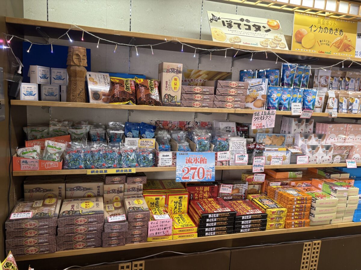 網走「丸北北都」の店内。手前の棚には地酒が並び、その奥には網走名物の「流氷飴」や、北海道の銘菓・箱菓子が棚いっぱいに陳列されている。海産物のみならず、網走ならではのスイーツや定番の北海道土産が豊富に揃う様子。
Inside Marukitahokuto in Abashiri, featuring local sake in the foreground. The background shelves are filled with Abashiri's famous "Ryuhyo Ame" (Drift Ice Candy) and various Hokkaido boxed sweets. It highlights the shop's extensive selection of both seafood and local confections.