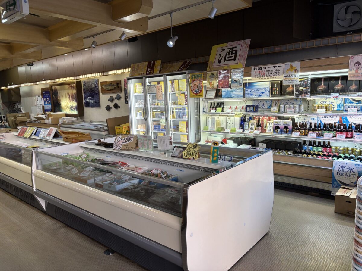 網走「丸北北都」の店内。手前の冷蔵ケースにはイクラや珍味などの加工海産物が並び、奥の棚には地酒などのアルコール類が豊富に陳列されている。上部にはカニの地方発送を案内する黄色い札が多数掲げられており、お土産選びに最適な活気ある店内の様子。
Inside Marukitahokuto in Abashiri, featuring a refrigerated display of processed seafood like salmon roe (ikura) in the foreground. The background shelves are stocked with various alcoholic beverages, including local Japanese sake. Yellow signs for crab shipping orders hang from the ceiling, showing a lively market scene.