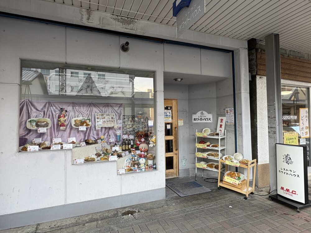 網走市の人気洋食店「レストラン ホワイトハウス」の外観。店頭のショーケースにはステーキやハンバーグなどの豊富な食品サンプルが並び、入り口横のラックにもおすすめメニューが掲示されている。 Exterior of "Restaurant White House" in Abashiri, featuring a glass showcase filled with various food samples of steaks, hamburgers, and seafood dishes.