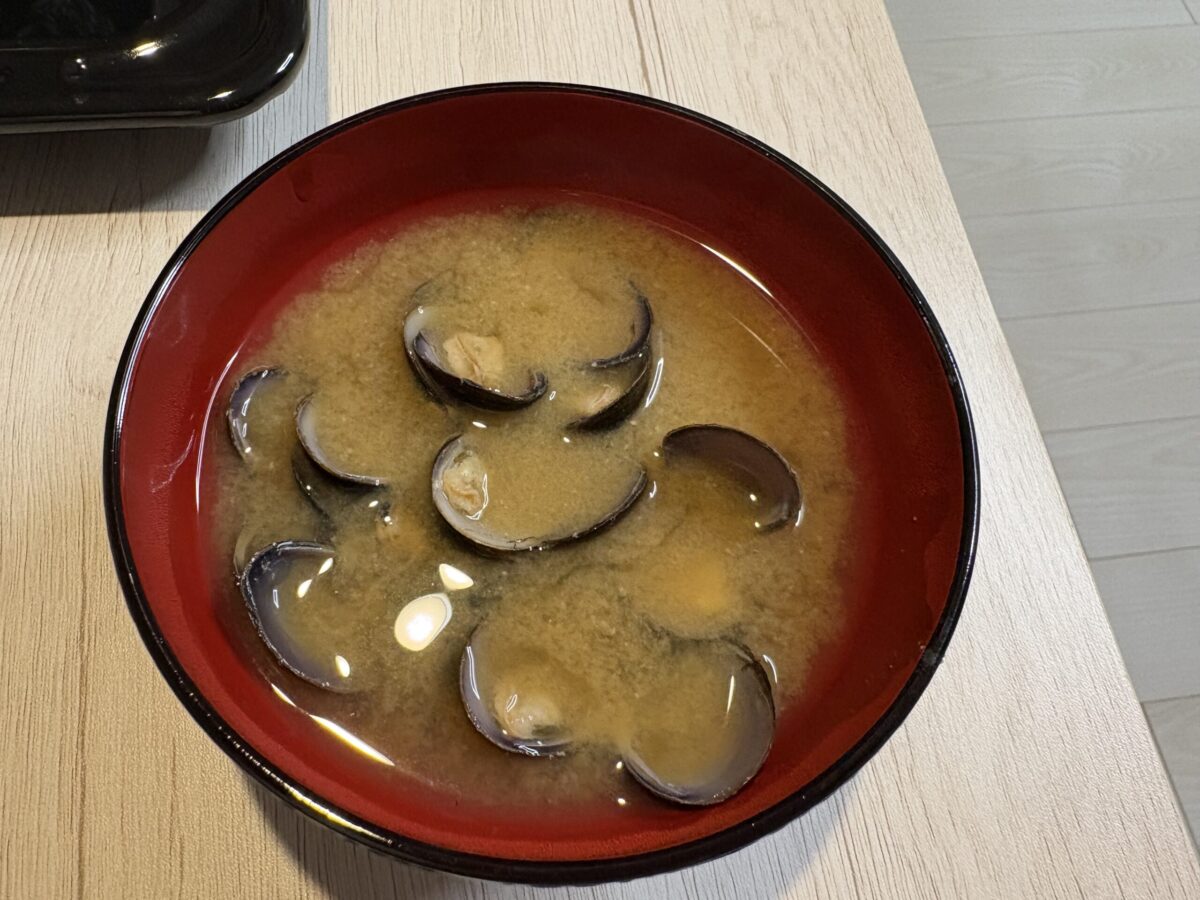 網走湖産の大きな「しじみ」をふんだんに使ったお味噌汁。お椀の中に黒く光る大粒のしじみが重なり合い、その濃厚なエキスが溶け出した温かい汁物の様子。
A bowl of steaming miso soup made with large Shijimi clams harvested from Lake Abashiri. The image shows the generous amount of dark, plump clams in a rich, flavorful broth.