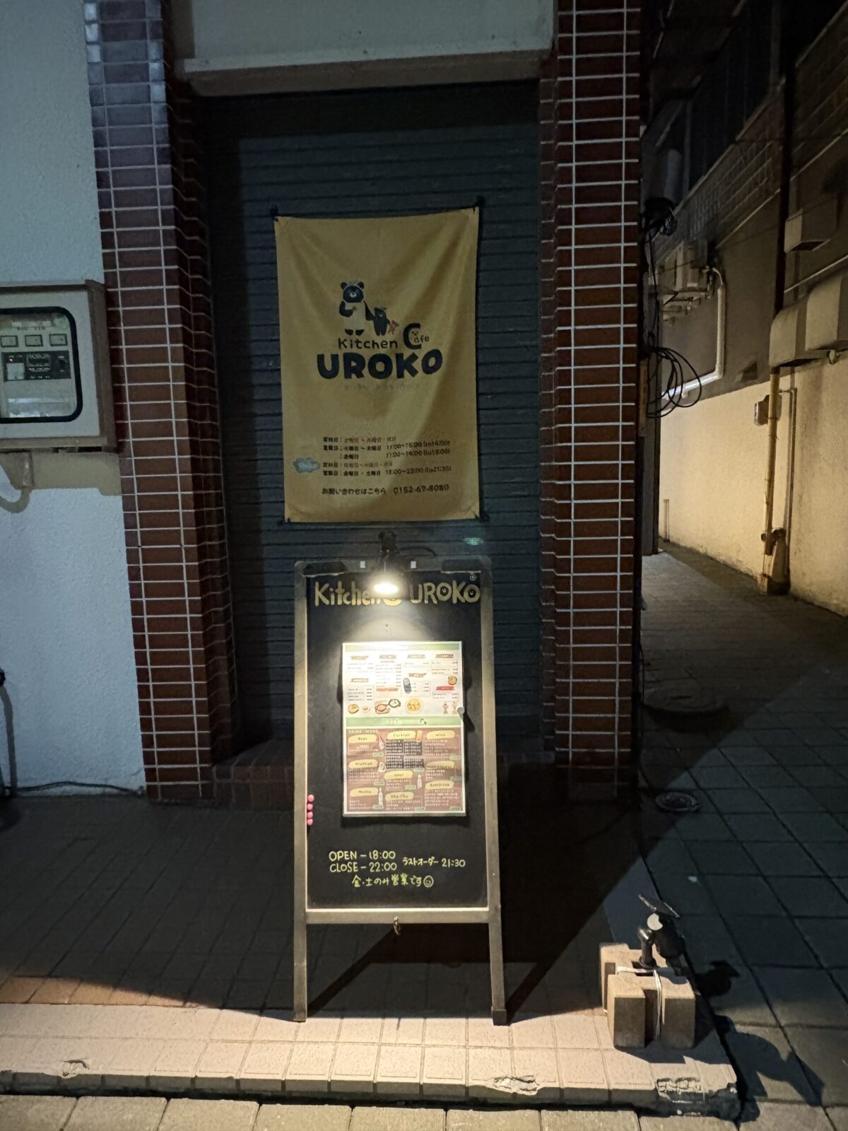 夜の「UROKO」路地入り口。シャッターの前に下げられた黄色い「Kitchen & Cafe UROKO」の暖簾が、スポットライトに照らされたメニュー看板と共に、通行人に路地の奥にお店があることを知らせている。
The alley entrance to "UROKO" in Abashiri at night. A yellow curtain for "Kitchen & Cafe UROKO" hangs above a spotlighted menu board, inviting passersby to explore the hidden alley.