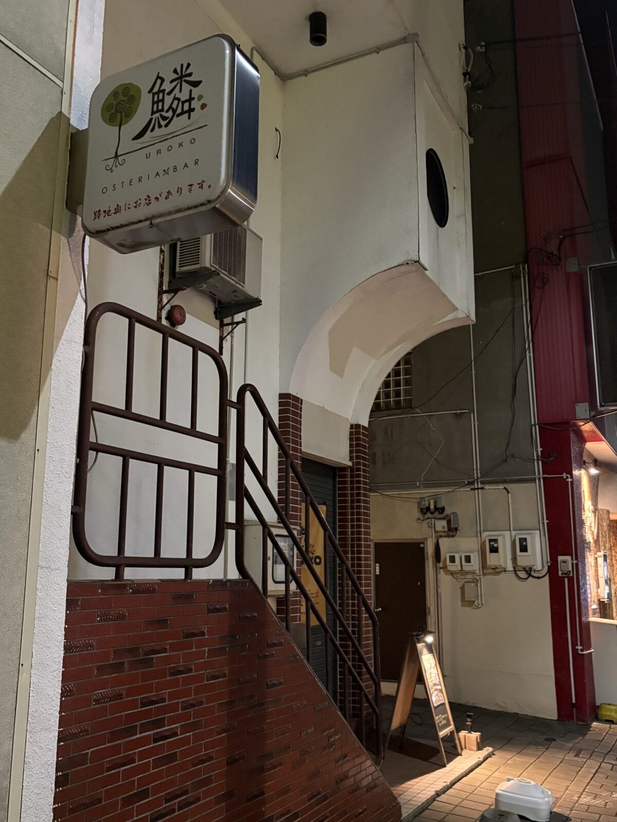 網走の繁華街にある「OSTERIA & BAR UROKO」の路地入り口看板。「鱗」という文字と木のイラストが描かれた白いキューブ型の看板の下に「路地奥にお店があります」と案内されている。
A street-side cube sign for "OSTERIA & BAR UROKO" in Abashiri. Below the sign, which features the Japanese character for "scale" (鱗), it directs customers: "The shop is located at the end of the alley."