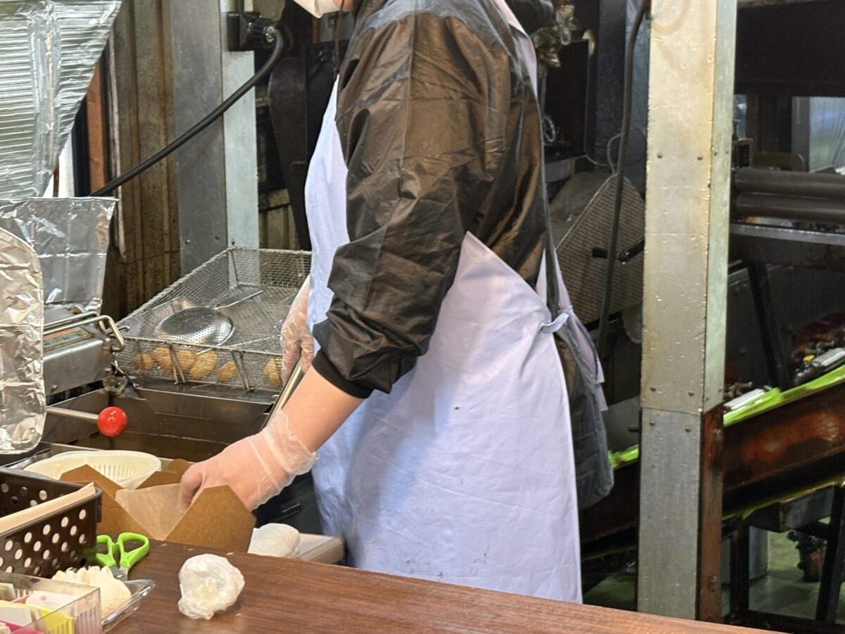 網走「横山蒲鉾店」の厨房で、職人が手際よくかまぼこを揚げている様子。フライヤーの横で、揚げたての黄金色のかまぼこを箱に詰める作業工程が写っている。
A scene inside the kitchen of Yokoyama Kamaboko in Abashiri, showing a staff member carefully preparing freshly fried fish cakes. The image captures the authentic local production process of golden-brown kamaboko.