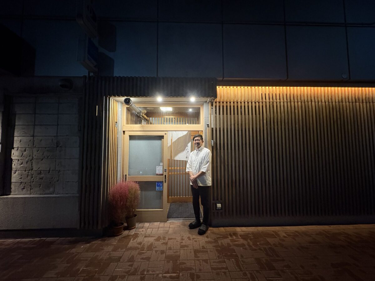 網走「鮨 やまし田」の店主（大将）が、清涼感のある白い暖簾が掛かった店門の前に立つ姿。職人の風格漂う白衣姿と、モダンな黒格子戸が調和した、お店の信頼感とおもてなしの心を感じさせる一枚。
The master chef and owner of Sushi Yamashita standing at the entrance of his restaurant in Abashiri. The crisp white Noren and modern black lattice door create a dignified atmosphere, welcoming guests for a premium dining experience.