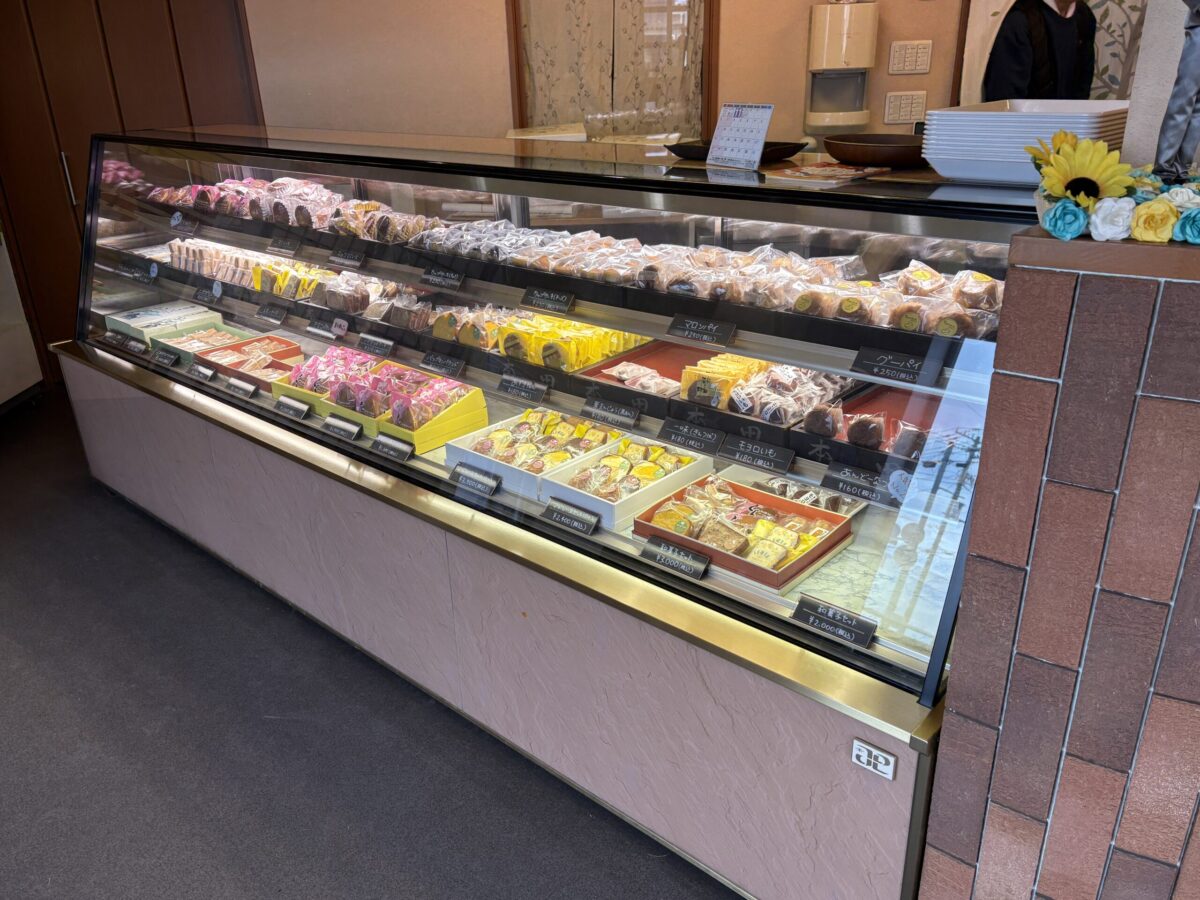 「本田菓子舗」の焼き菓子と和菓子のショーケース。贈答用の詰め合わせセットや、個包装されたマロンパイ、あんバタなどの地元の銘菓が綺麗に陳列されている。
A display case featuring baked goods and traditional Japanese sweets at Honda Kashiho. It shows various gift boxes and individually wrapped treats like chestnut pies and "An-バター."