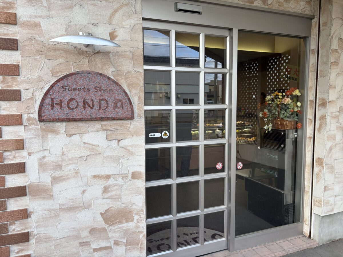 「Sweets Shop HONDA」と刻まれたアーチ型の石造り看板が目を引く、本田菓子舗の店舗入り口。ガラス張りの自動ドアからは店内の明るい雰囲気が伺える。
The entrance of Honda Kashiho featuring an arched stone sign that reads "Sweets Shop HONDA." The glass automatic door provides a glimpse into the welcoming interior.