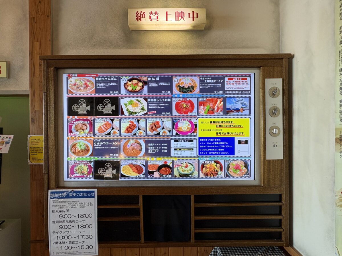 映画館の掲示板を模した「キネマ館」のメニュー一覧。「絶賛上映中」の看板の下に、網走ちゃんぽん、カニ飯、オホーツク干貝柱塩ラーメンなど、網走ならではの多彩な料理がフィルム風の枠に並んでいる。
A restaurant menu board designed like a retro cinema display at Kinemakan. It features local dishes such as Abashiri Chanpon and Crab Rice Bowl framed in film-strip style, with a "Now Showing" sign at the top.