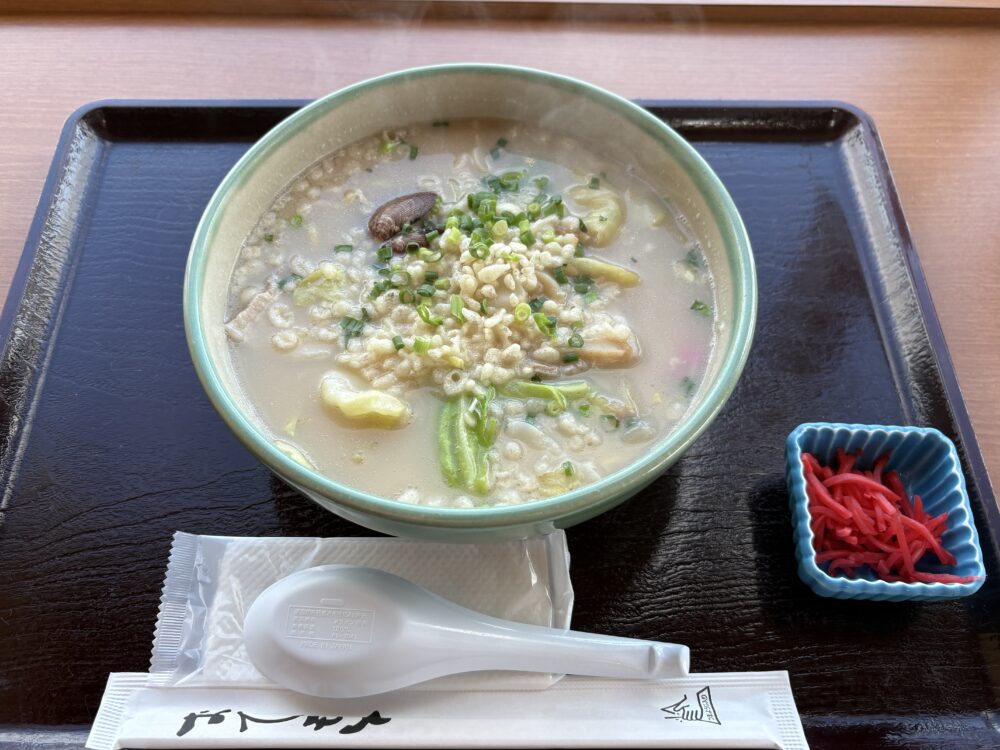 道の駅「流氷街道網走」のキネマ館で提供される、網走ちゃんぽんのお膳。白濁したまろやかなスープに、たっぷりの天かす、野菜、アサリが入り、別皿で紅ショウガが添えられている。 A meal tray featuring Abashiri Chanpon, a local noodle specialty at Kinemakan. The bowl contains a creamy white broth with tempura bits, vegetables, and clams, served with a side of red pickled ginger.