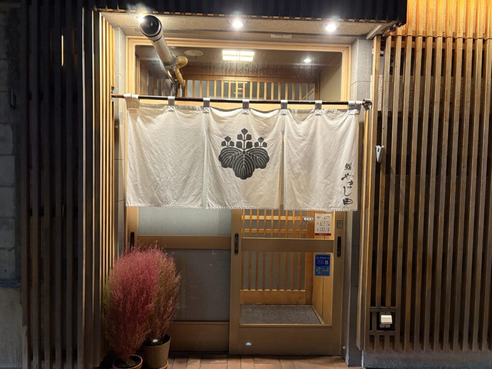 網走市の本格寿司店「鮨 やまし田」の入り口。シックな黒の格子戸に、清潔感のある真っ白な暖簾が掛かったモダンで高級感漂う店構え。夜の網走で静かにゲストを迎える隠れ家のような佇まい。 The elegant entrance of Sushi Yamashita in Abashiri. It features a modern black lattice door and a crisp white Noren (curtain), creating a sophisticated and welcoming atmosphere for a high-end sushi dining experience.