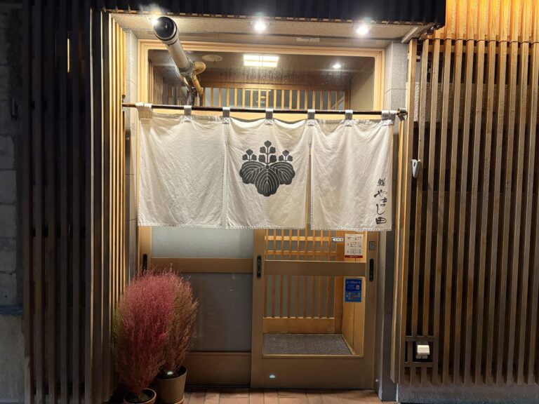 網走市の本格寿司店「鮨 やまし田」の入り口。シックな黒の格子戸に、清潔感のある真っ白な暖簾が掛かったモダンで高級感漂う店構え。夜の網走で静かにゲストを迎える隠れ家のような佇まい。 The elegant entrance of Sushi Yamashita in Abashiri. It features a modern black lattice door and a crisp white Noren (curtain), creating a sophisticated and welcoming atmosphere for a high-end sushi dining experience.