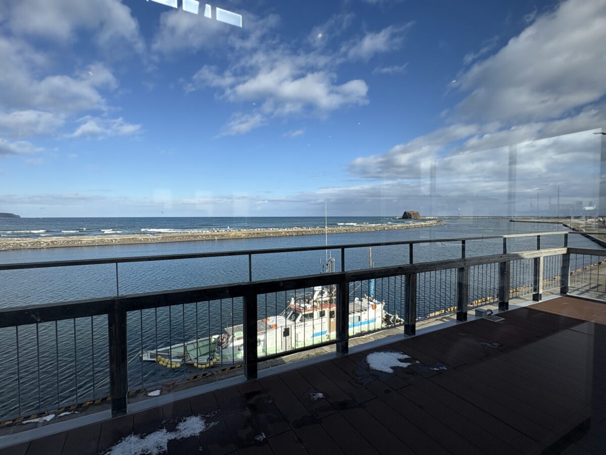 道の駅「流氷街道網走」の2階にあるレストラン・キネマ館から望む、網走港とオホーツク海の青い景色。手前には漁船が停泊し、遠くに防波堤が見える。
A scenic view of Abashiri Port and the Sea of Okhotsk from Kinemakan restaurant at the Abashiri Roadside Station, featuring a fishing boat docked at the pier under a blue sky with white clouds.