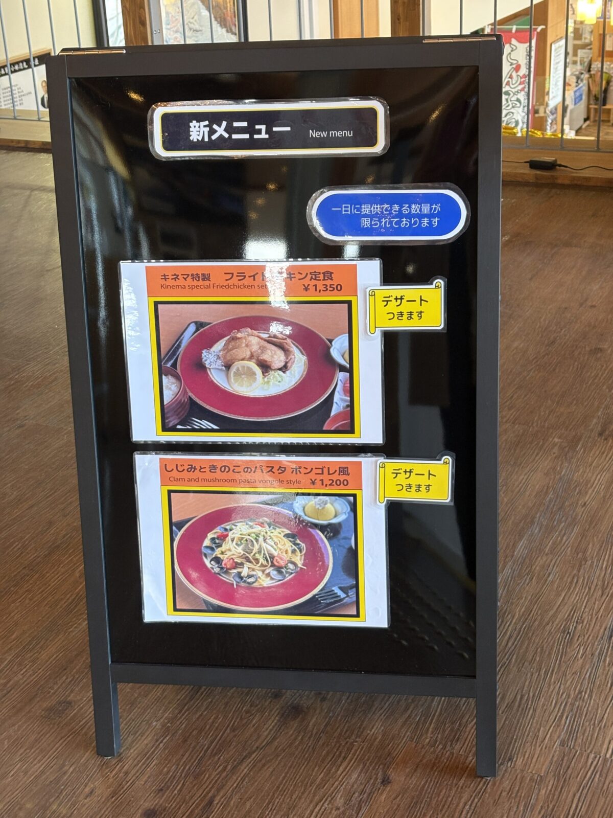 レストラン「キネマ館」の入り口に設置された新メニューの立て看板。「キネマ特製フライドチキン定食」と「しじみときのことパスタ ボンゴレ風」が、デザート付きで紹介されている。
An outdoor A-frame sign showcasing new menu items at Kinemakan: Special Fried Chicken Set and Clam & Mushroom Pasta Vongole style, both served with dessert.