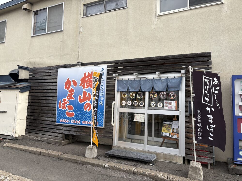 網走市の老舗「横山蒲鉾店」の店舗入り口。青い背景に赤い文字で「横山の立かまぼこ」と書かれた大きな看板と、「あばしり揚げたてかまぼこ」の黒いのぼりが立っている、木目調の外壁の店構え。 The storefront of Yokoyama Kamaboko, a long-established fish cake shop in Abashiri. It features a prominent blue and red sign and a black banner advertising "freshly fried Abashiri kamaboko."