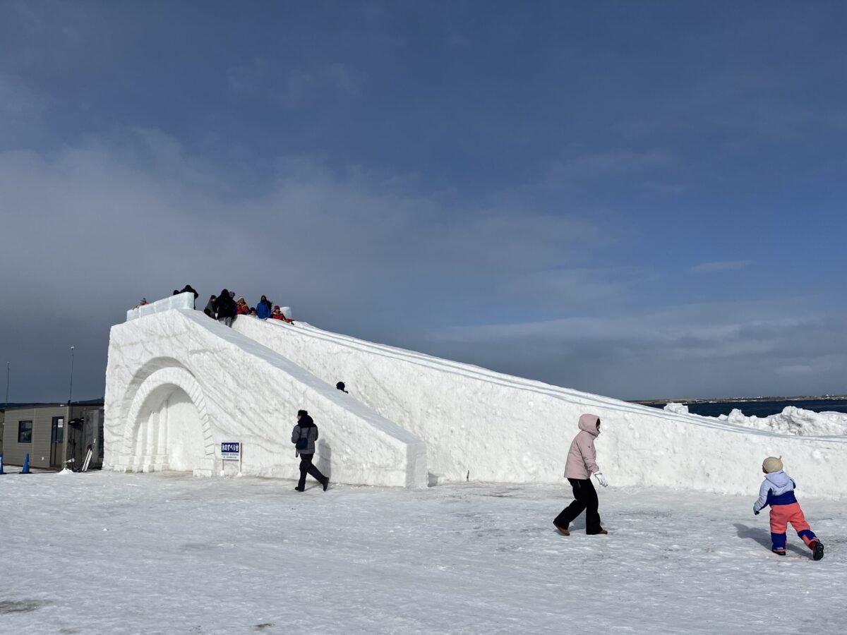 快晴の下、北海道のあばしりオホーツク流氷まつりに設置された、子供や家族向けの巨大な雪の滑り台。
A massive snow slide for children and families at the Abashiri Okhotsk Drift Ice Festival in Hokkaido, under a clear blue sky.