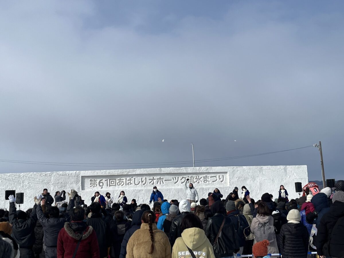 第61回あばしりオホーツク流氷まつりの文字が刻まれた巨大な雪のステージで、冬服を着た大勢の観客がイベントを観覧している様子 A large crowd of people in winter coats watching an event at a massive snow stage engraved with the name of the 61st Abashiri Okhotsk Drift Ice Festival.