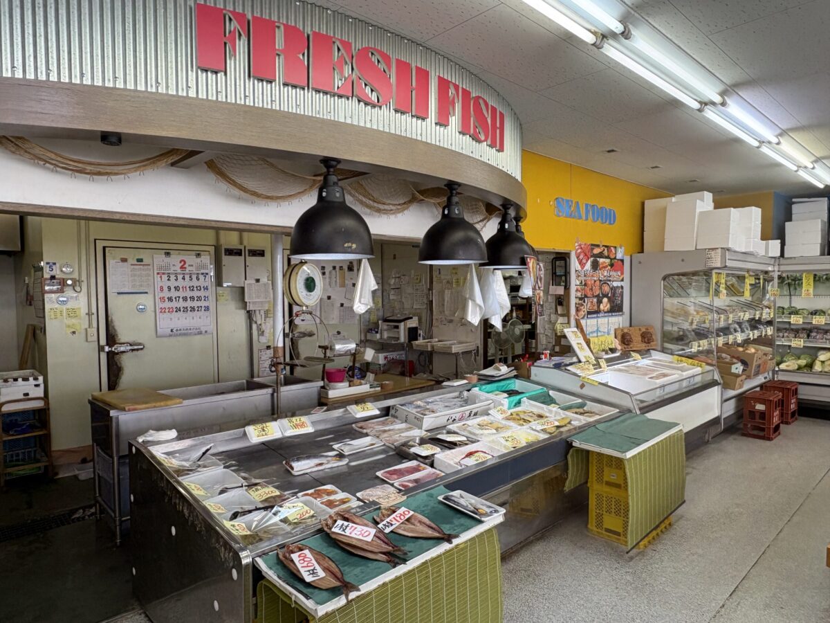 網走「はせがわ」店内の鮮魚コーナー。「FRESH FISH」の赤い看板の下に、地元の新鮮な魚介類や干物が並ぶ様子。
nside Hasegawa Store in Abashiri, showing the "FRESH FISH" section with a variety of local seafood and dried fish displayed on refrigerated counters.