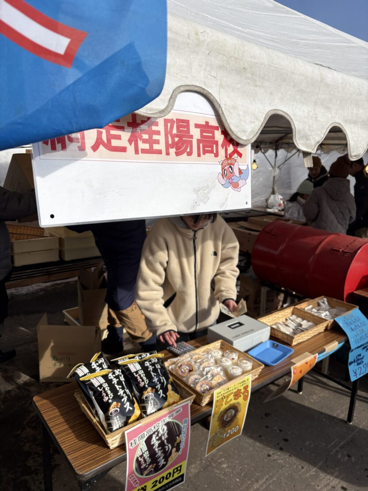 網走桂陽高校の看板の下で接客する生徒の様子。カウンターには「あばしりポテトチップス」や「ぽてまる」といった地元のスナックが並んでいる
A student serving customers at a festival stall under a sign for Abashiri Keiyo High School, featuring local snacks like Abashiri Potato Chips and Potemaru on the counter.