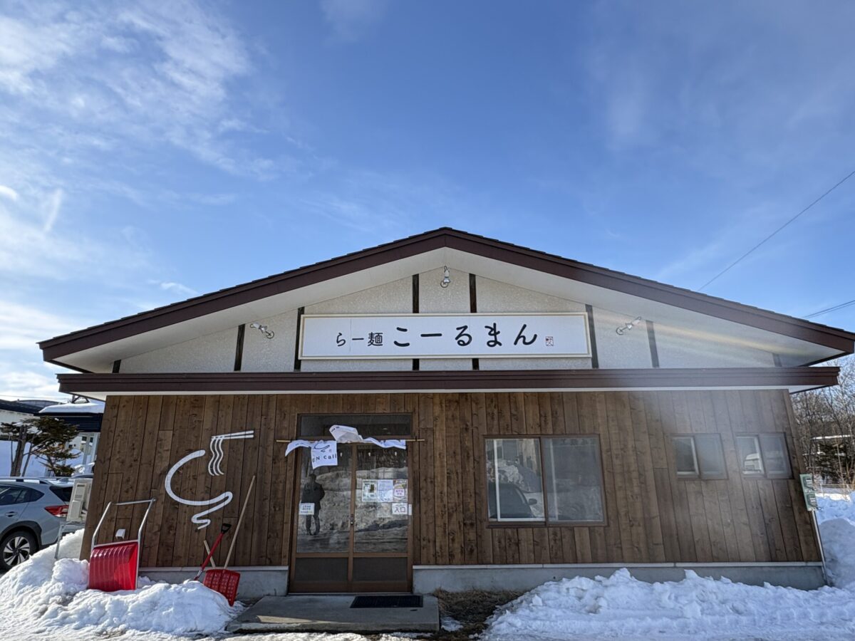 「らー麺 こーるまん」の店舗外観。木目調の壁に三角屋根の平屋建てで、入り口横には麺をリフトアップしたユニークなロゴが描かれている。建物の周りには高く積もった雪があり、冬の網走らしい風景。
The exterior of "RAMEN callman" restaurant in Abashiri. A one-story building with brown wooden siding and a gabled roof. A stylized ramen noodle logo is painted on the wall. The ground is covered with piles of white snow against a clear blue sky.