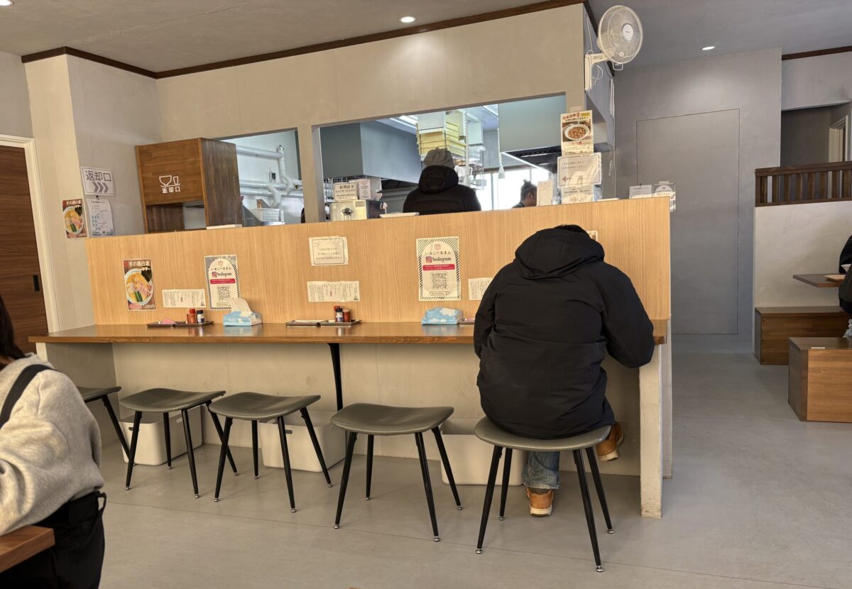 「らー麺 こーるまん」の店内。木目調のカウンター席が並び、客が背を向けて食事をしている。カウンターには調味料、ティッシュ、InstagramのQRコードが置かれている。奥の調理場との間には「返却口」が設けられており、全体的に清潔でモダンな雰囲気。
The interior of "Ramen Callman," showing a row of wooden counter seats with customers dining. Seasonings, tissues, and an Instagram QR code are on the counter. A dish return window is visible in the background leading to the kitchen. The overall atmosphere is clean and modern.