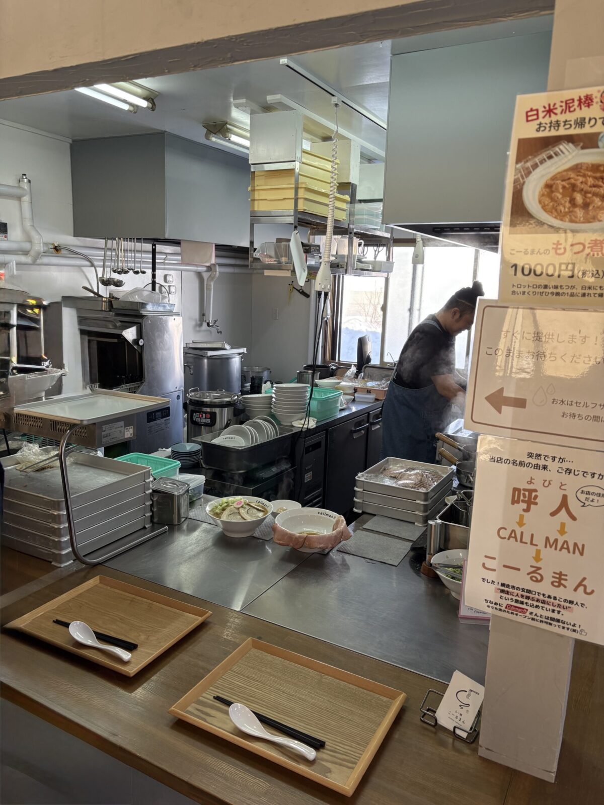 網走市のラーメン店「らー麺 こーるまん」のオープンキッチン（調理場）。店主がラーメンを調理しており、カウンターには提供待ちのラーメン2杯が置かれている。右側の仕切り板には、店名の由来が「呼人（よびと）→CALL MAN→こーるまん」であることを説明するポスターや、テイクアウトメニュー「白米泥棒のもつ煮」のポスターが掲示されている
he open kitchen area of "Ramen Callman" in Abashiri. The owner is preparing ramen, and two bowls of ramen are waiting on the counter. Posters on the partition board explain the restaurant's name origin ("Yobito" area -> CALL MAN -> Callman) and advertise a takeout "Motsu Stew" dish.