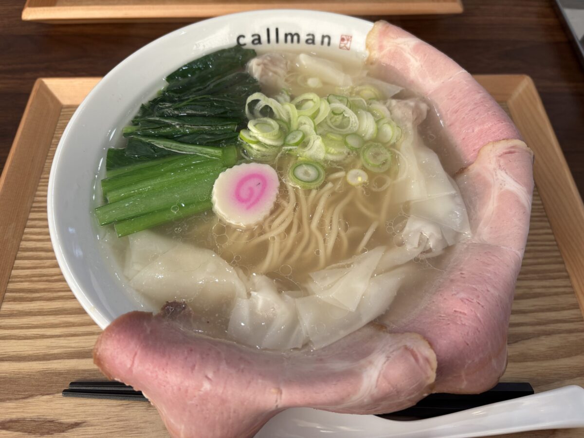 網走市の「らー麺 こーるまん」で提供される、澄んだ塩スープの「コールマン塩ラーメン」。白い丼の縁を、大きなピンク色のレアチャーシュー2枚が覆うようにトッピングされている。麺の上には、ワンタン、たっぷりのほうれん草、刻みネギ、そしてナルトが乗っている。丼には「callman」のロゴが見える。
A hearty bowl of "Callman Shio Ramen" from "Ramen Callman" in Abashiri, Hokkaido. It features a clear, savory salt broth. Two large slices of pink, thinly sliced rare chashu pork are draped over the rim of the white bowl. The ramen is topped with several wontons, generous portions of spinach, finely chopped green onions, and a pink-and-white fish cake (naruto). The bowl has the restaurant's logo "callman" printed on it.