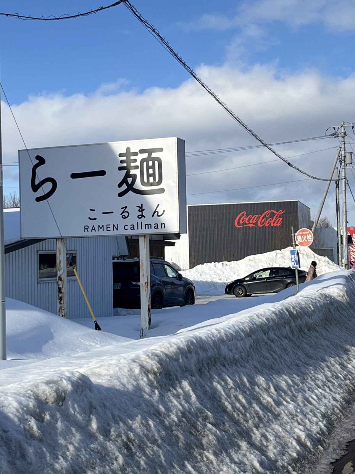 網走市のラーメン店「らー麺 こーるまん」の屋外看板。白地に黒文字で店名が書かれ、背景には積雪した道路とコカ・コーラの赤いロゴが入った建物、青空が広がっている。
A roadside sign for "RAMEN callman" (Ramen Callman) in Abashiri, Hokkaido. The sign features bold black Japanese characters on a white background. The surrounding landscape is covered in thick snow under a bright blue sky, with a Coca-Cola building visible in the distance.