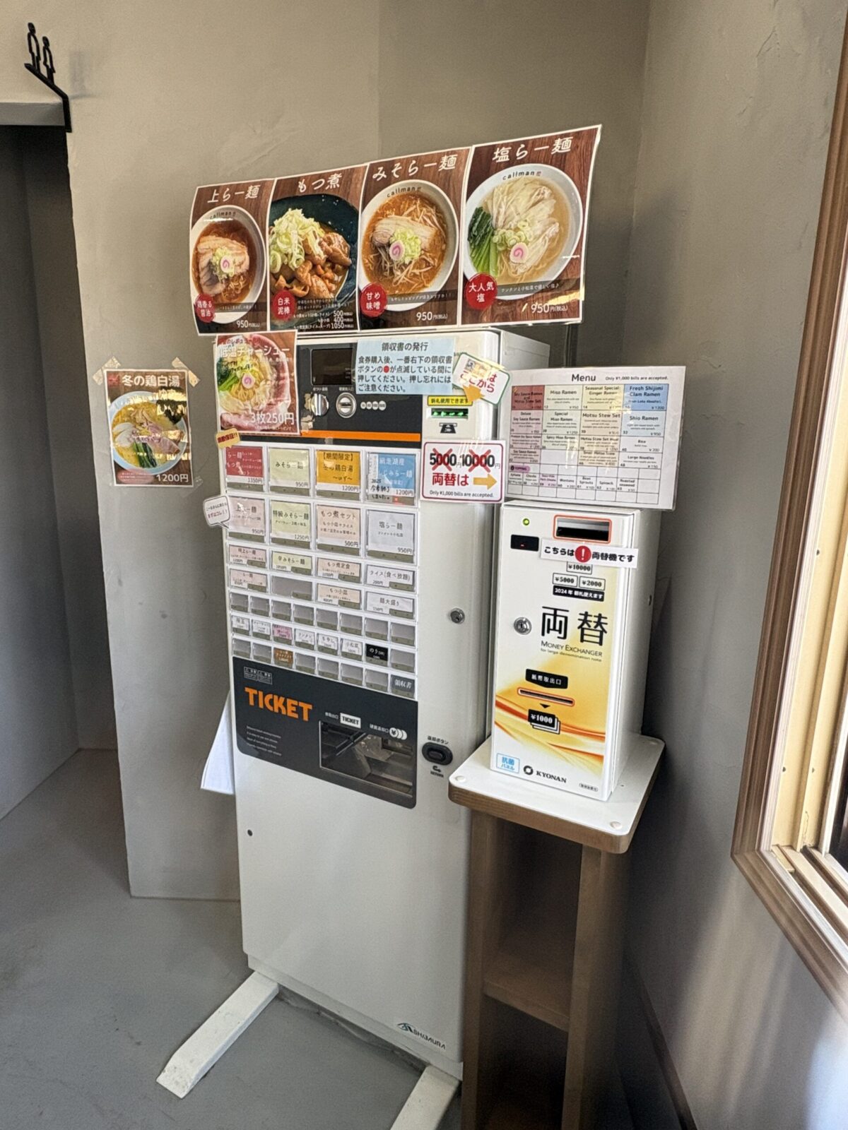 網走市の「らー麺 こーるまん」の券売機と両替機。券売機の上には「上らー麺」「もつ煮」「みそらー麺」「塩らー麺」の大きな写真メニューが掲示されている。横には英語表記のメニューリストや、冬季限定の「冬の鶏白湯」のポスターも貼られており、千円札以外も使えるよう高額紙幣の両替機が併設されている。
A ticket vending machine and money changer at "Ramen Callman" in Abashiri. Large photos above the machine show popular dishes like "Special Soy Sauce Ramen," "Motsu Stew," "Miso Ramen," and "Shio Ramen." An English menu and a seasonal "Winter Chicken Paitan" poster are also visible. A separate money changer is placed next to the machine for 5,000 and 10,000 yen bills.