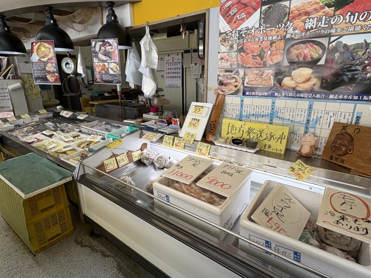 網走の商店「はせがわ」の店内ショーケース。手前にはタラバガニ、ズワイガニ、毛ガニの発泡スチロール箱、奥には新鮮な魚介類が並ぶ。壁には「網走の旬」を紹介するポスターと「地方発送承り中」の案内が掲示されている。
Inside the Hasegawa Store in Abashiri, showing a display case filled with local Hokkaido seafood like King Crab, Snow Crab, and Horsehair Crab, alongside a variety of fresh fish. Promotional posters for Abashiri's seasonal seafood are visible in the background.