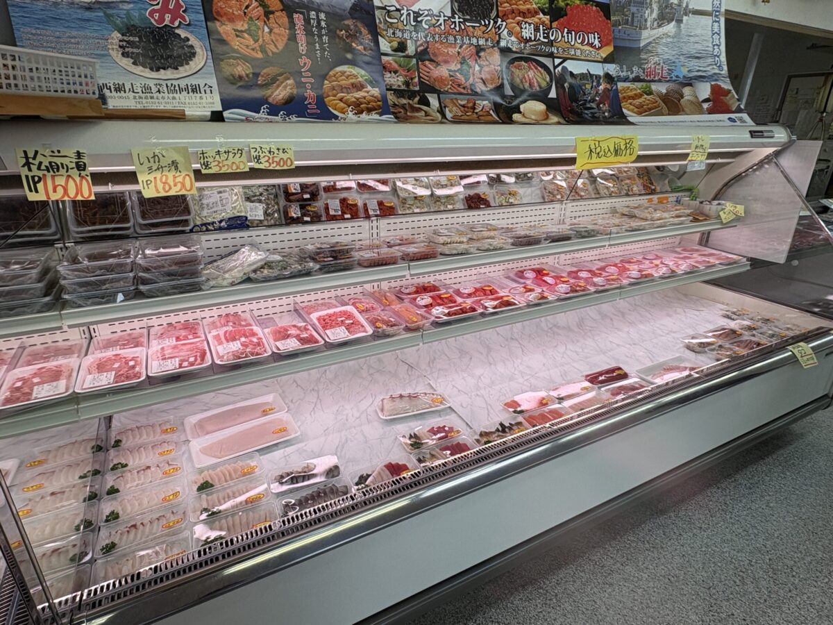 網走「はせがわ」の精肉・加工品コーナー。ジンギスカン用のお肉や精肉パックが並ぶショーケース、上段には松前漬やつぶわさびなどの地元の味が並ぶ。
Meat and delicatessen section at Hasegawa Store in Abashiri, Hokkaido. The refrigerated display shows various cuts of meat, including Jingisukan (grilled mutton), alongside local specialties like Matsumaezuke (pickled seafood).