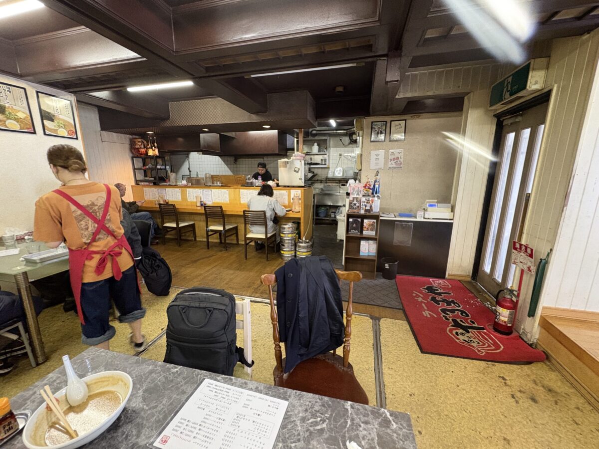 「ラーメンだるまや 網走店」の店内の様子。木目調のカウンター席、テーブル席があり、スタッフが忙しく立ち働く活気ある雰囲気。入り口には赤い店名入りのマットが敷かれ、手前のテーブルには食べ終わりのラーメンの器とメニューが置かれている。
The interior of "Ramen Darumaya" in Abashiri, Hokkaido. It shows a cozy atmosphere with wooden counter seats, a busy staff member in a red apron, and a red welcome mat at the entrance. A finished bowl of ramen and a menu are visible in the foreground.