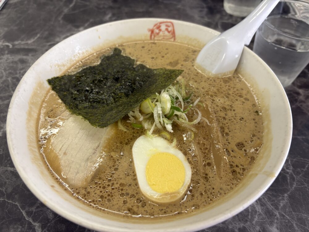「ラーメンだるまや 網走店」の一番人気メニュー、どろラーメン。豚骨ベースの濃厚でクリーミーな茶褐色のスープが泡立ち、トッピングには大きな海苔、チャーシュー、ゆで卵、刻みネギが添えられている。だるまのロゴが入った白い器に盛られた一杯。 The signature "Doro Ramen" at Ramen Darumaya in Abashiri, Hokkaido. It features a rich, creamy, and frothy pork bone soy sauce broth. The bowl is topped with a large sheet of Nori seaweed, sliced chashu pork, a boiled egg, and fresh green onions.