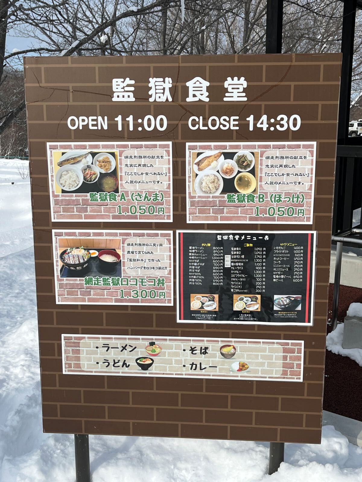 博物館 網走監獄「監獄食堂」の屋外営業案内看板。現在の刑務所の食事を再現した「監獄食」の文字が目立つ入り口の風景
Outdoor welcome sign of the Abashiri Prison Museum cafeteria, highlighting the "Prison Meal" available inside.
