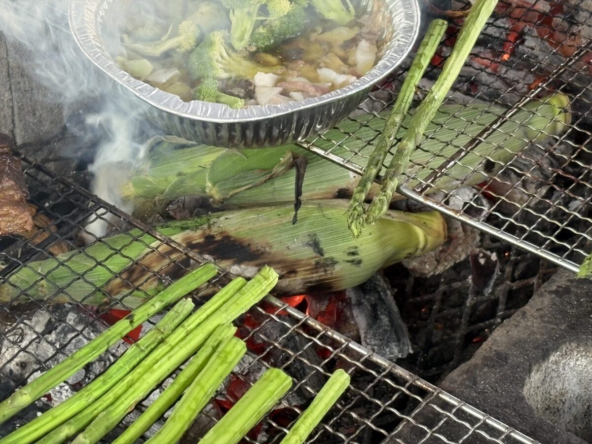 網走の夏、屋外で楽しむ炭火バーベキュー。網の上で香ばしく焼かれる皮付きのトウモロコシと鮮やかな緑のアスパラガス、アルミ皿で煮えるブロッコリーのアヒージョ。
A charcoal BBQ in Abashiri during summer, featuring local seasonal vegetables. Fresh corn in husks and vibrant green asparagus are being grilled on a wire rack next to a foil pan of broccoli ajillo.